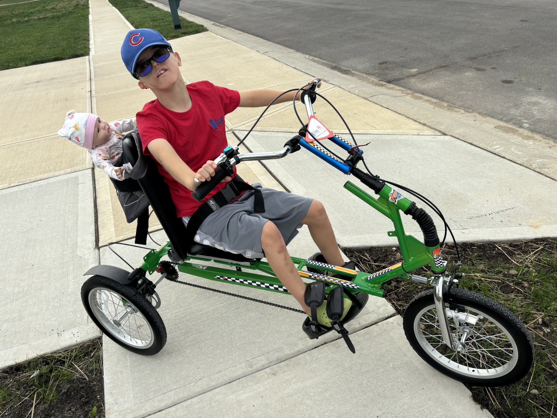 A boy rides his green adaptive bike with a doll in the back and looks up at the camera with sunglasses on. 