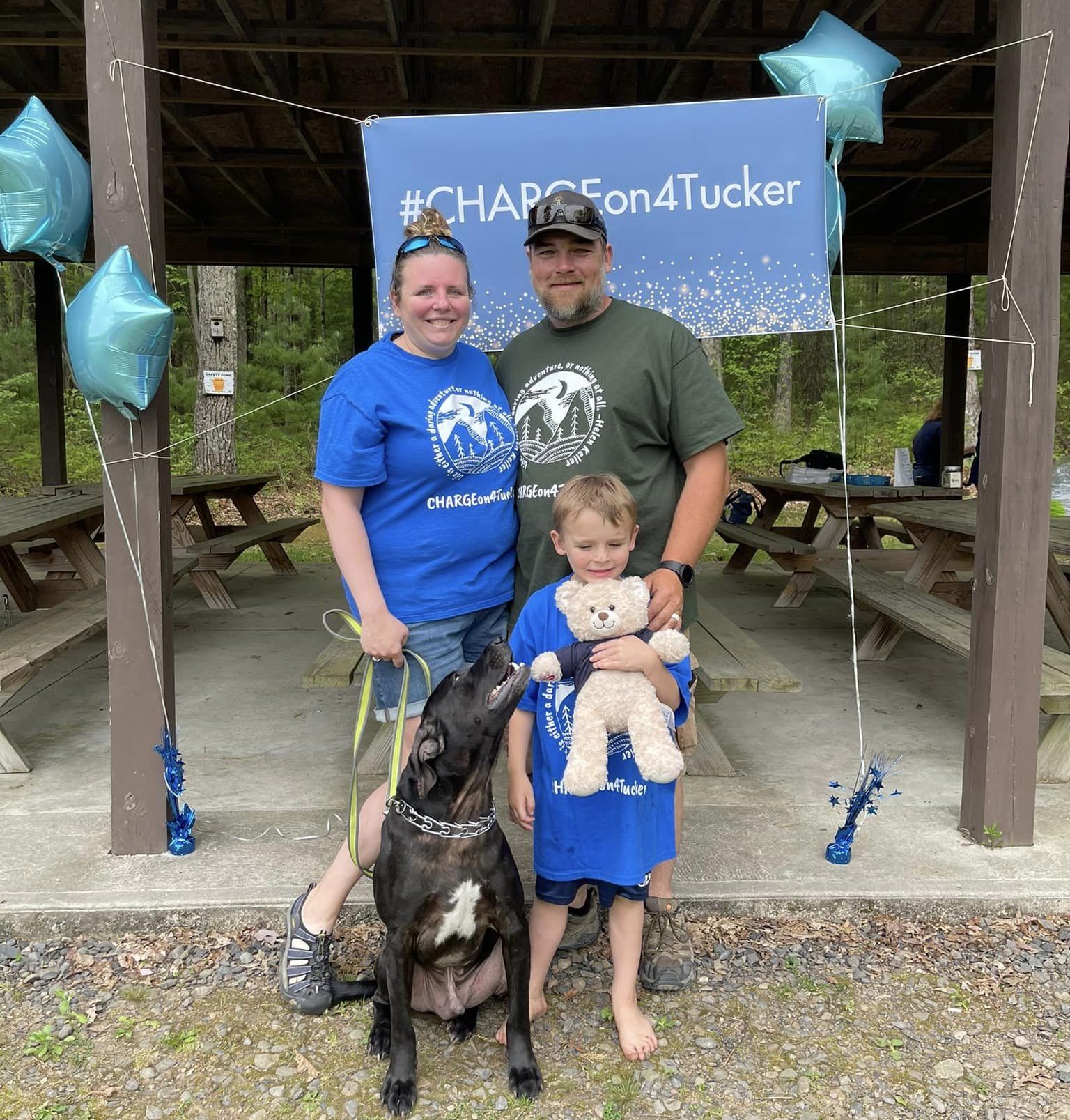Parents stand in front of a banner that reads CHARGE on for Tucker. A young boy, Hunter, holds on to a teddy bear that contain his brother Tucker's ashes, as the family dog looks on. 