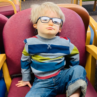 Young boy in a striped shirt and glasses sits in a chair.