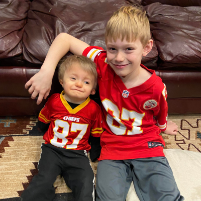 Two young brothers sit on the floor next to each other, wearing matching Kansas City Chiefs football jerseys. 