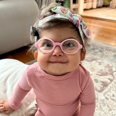 Adorable baby girl with a bow in her dark brown hair, in pink glasses, wearing a pink outfit, smiles at the camera.