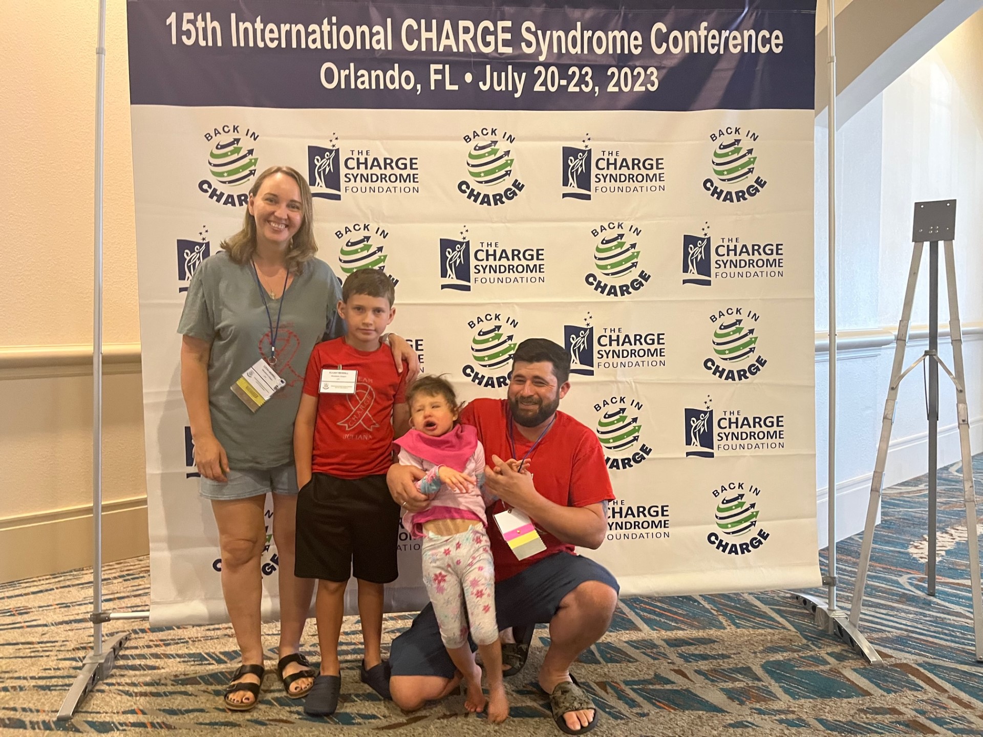 Family with two parents and two kids stands in front of the CHARGE Syndrome Foundation backdrop at Conference