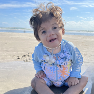 Baby boy with a trach sits on the beach. 