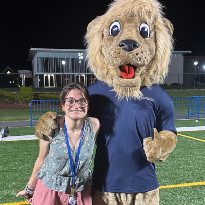 Happy teen girl with short brown hair and glasses poses with a lion mascot on a football field.