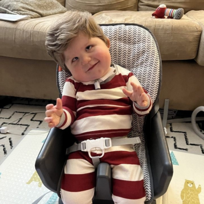 Baby boy with CHARGE syndrome has a trach, wears a striped outfit, and sits in a chair. 