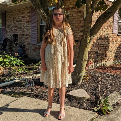 Girl with long blonde hair and glasses in a pretty yellow dress with sandals poses outside.