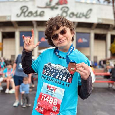 Young man in sunglasses poses outside the Rose Bowl stadium signing "I Love You", wearing a 5k Run bib. 