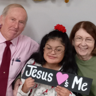 Photo of young girl with glasses holding a sign that says "Jesus Loves Me' between her parents. 