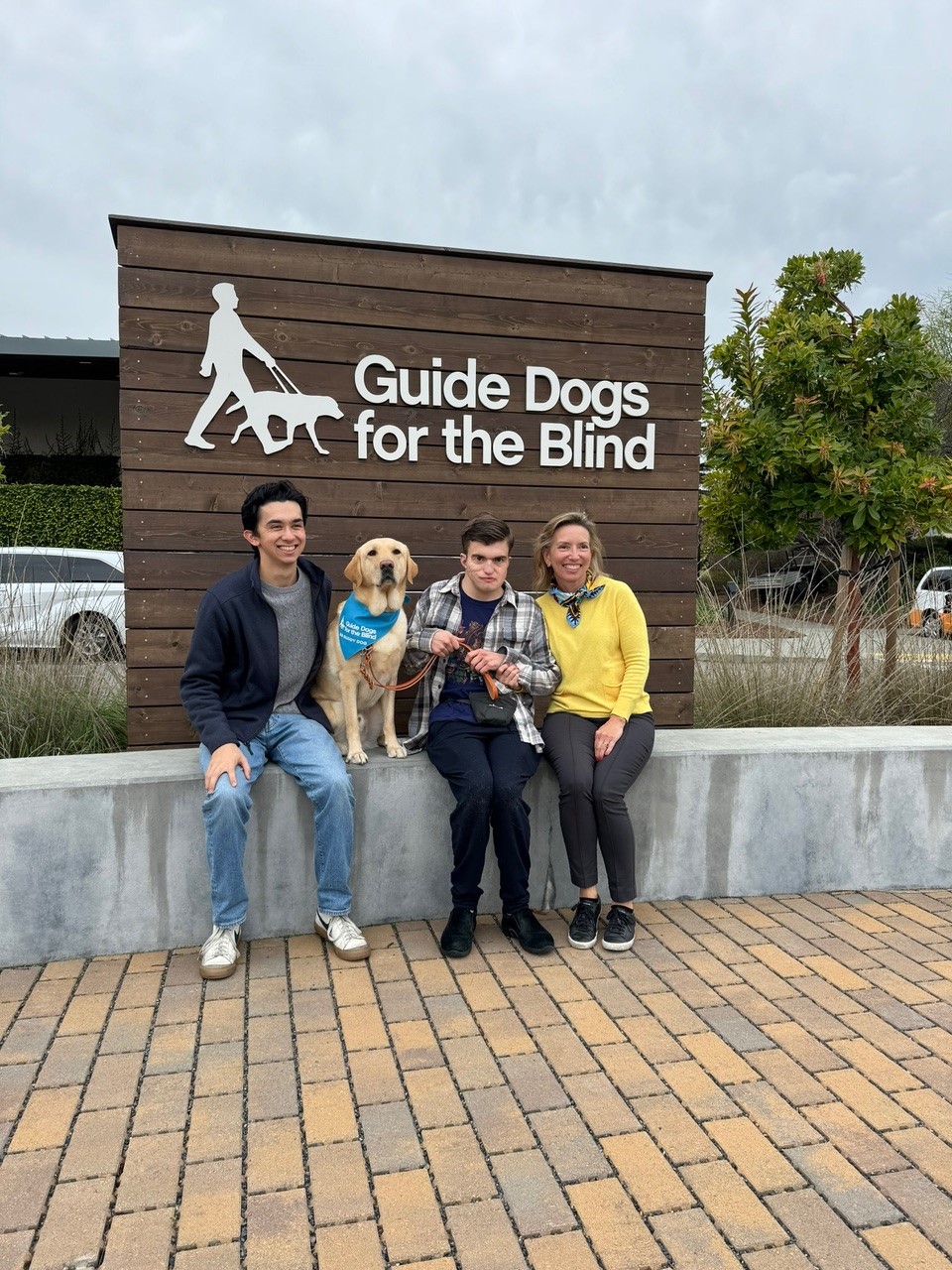 A young man sits with his golden retriever, and his family in front of a sign that reads Guide Dogs for the Blind