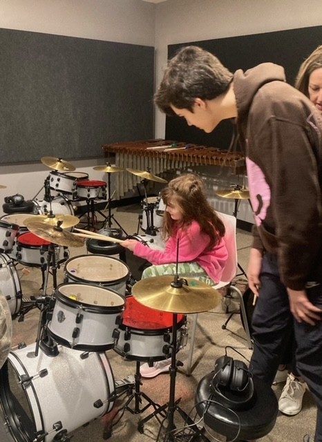 Adult looks on as little girl plays a drum set.