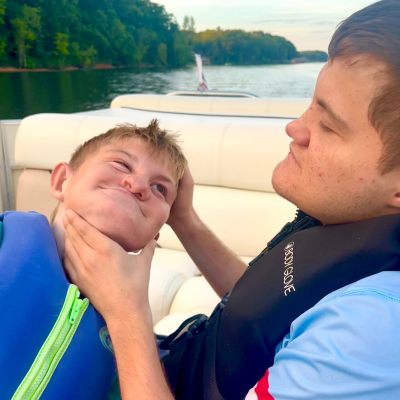 2 boys with CHARGE sit on a boat with life jackets on. One cradles the other's head