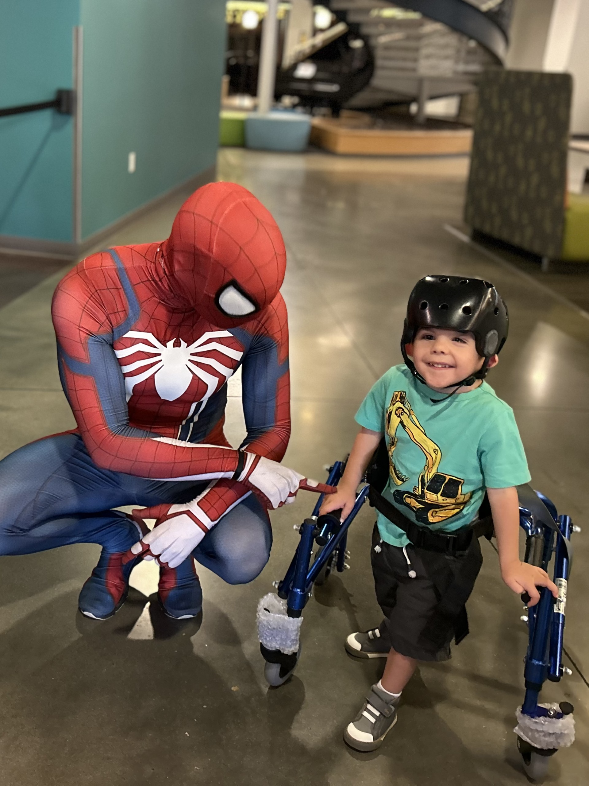 Young boy with a big smile using a walker and wearing a medical helmet poses with Spider Man. 