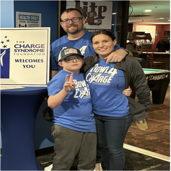 Family with Mom, Dad, and son pose by a sign for the CHARGE Syndrome Foundation. 