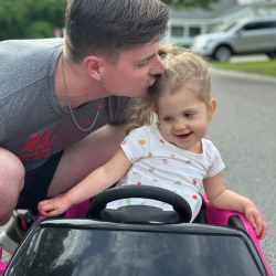 Father kisses his daughter's head as she rides in a toy car. 