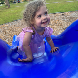 Young girl with CHARGE syndrome looks up smiling from the bottom of a slide