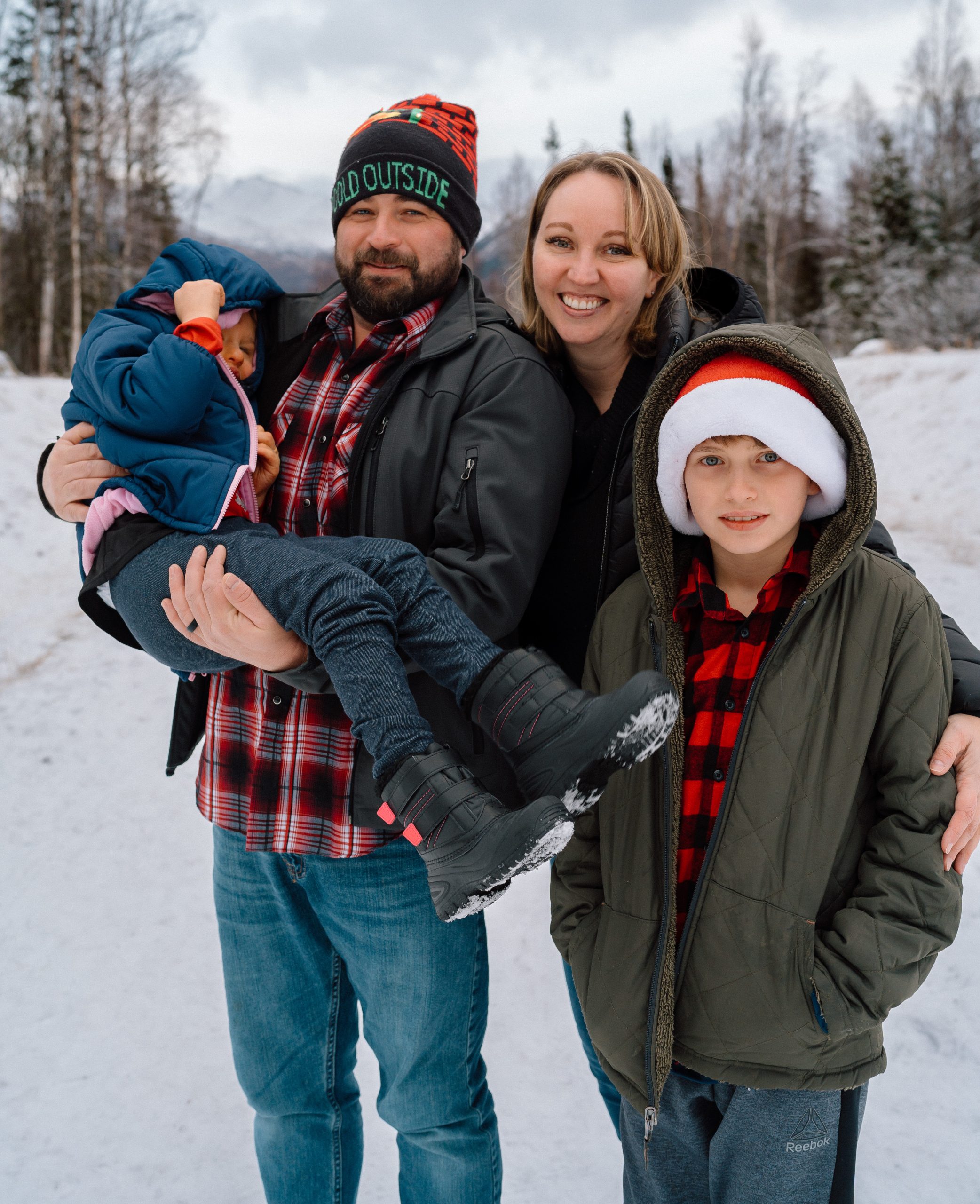 Family in winter coats standing in the snow. The father holds his daughter with CHARGE syndrome. Both the father and son wear santa hats.